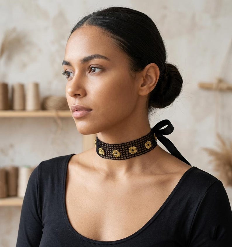 May include: A black choker necklace with a floral design. The choker is made of a mesh material and features gold-coloured flower accents. A black ribbon ties the necklace at the back. The person is wearing a black long-sleeve shirt.