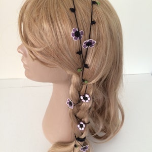 May include: A black hair accessory with purple and white crocheted flowers. The accessory is worn in a braid and around the head.
