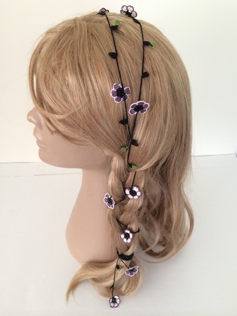 May include: A black hair accessory with purple and white crocheted flowers. The accessory is worn in a braid and around the head.