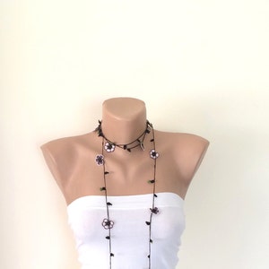 May include: A long black necklace with small white and purple flowers and green leaves. The necklace is layered and hangs down to the chest.