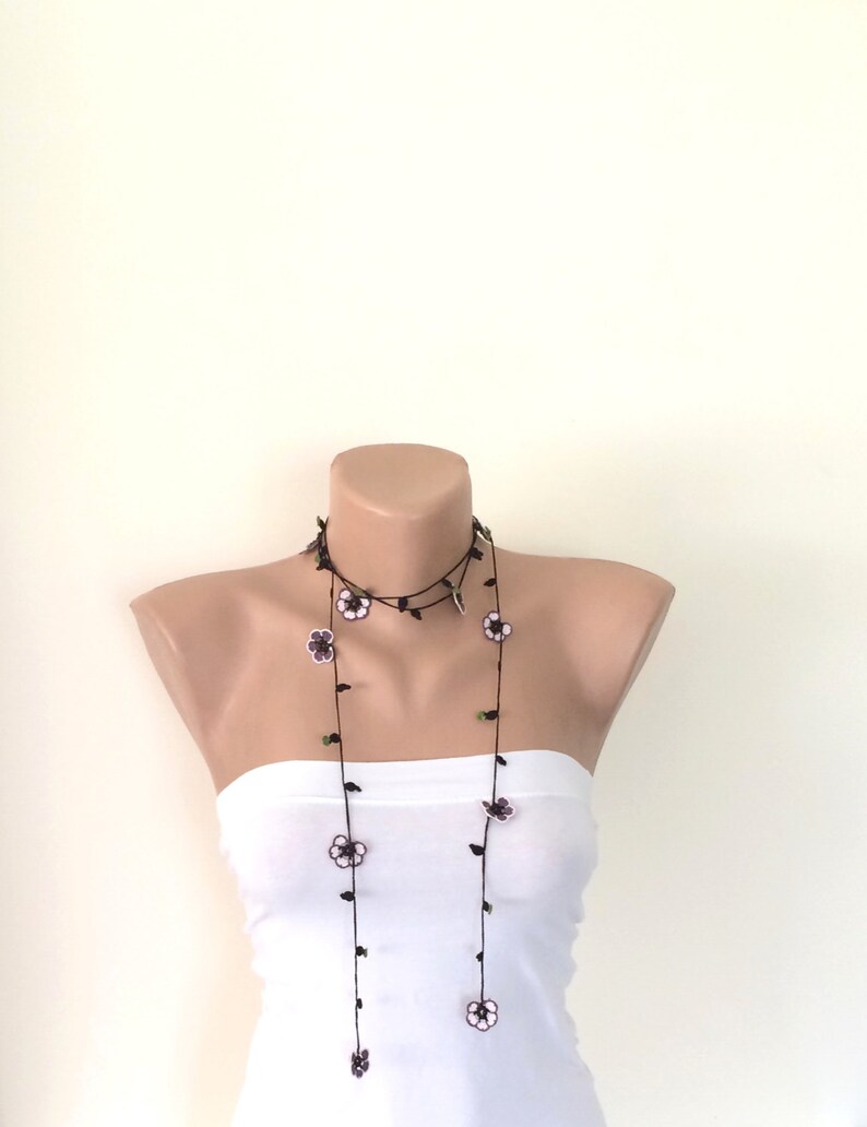 May include: A long black necklace with small white and purple flowers and green leaves. The necklace is layered and hangs down to the chest.