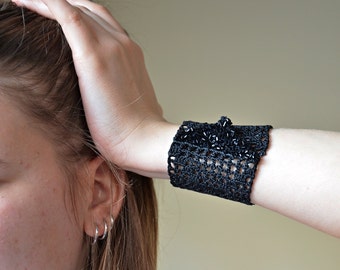 Puede incluir: Un brazalete de crochet negro con cuentas negras. El brazalete está en la muñeca de una persona.