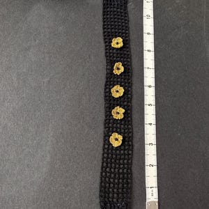 May include: Black crocheted headband with five gold-coloured flower accents. The headband measures approximately 30 cm in length. The black band has a velvet texture and is shown on a dark grey surface.