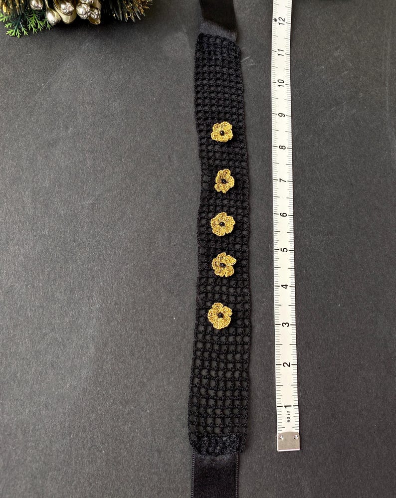 May include: Black crocheted headband with five gold-coloured flower accents. The headband measures approximately 30 cm in length. The black band has a velvet texture and is shown on a dark grey surface.
