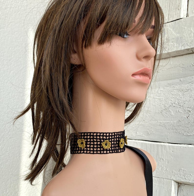 May include: Black crochet choker necklace with gold-coloured flower accents. The choker is worn around the neck and has a delicate, open weave design. The flowers are evenly spaced across the front of the choker.