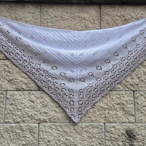 May include: A triangular white knit shawl featuring a central stockinette stitch section with a vertical spine, bordered by an intricate lace pattern along the edges. The textile displays a delicate, openwork design with repeating floral motifs.