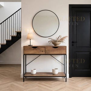 May include: A rectangular wooden console table with a black metal frame, two drawers, and a lower shelf. The table features a round wall mirror, a small table lamp, and a bowl with dried botanicals. Two white ceramic pots sit on the bottom shelf.