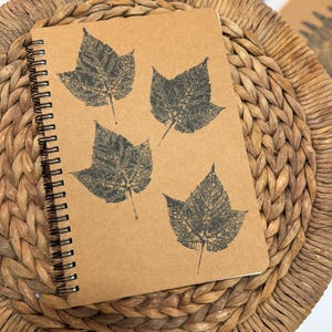 May include: A spiral-bound notebook with a light brown kraft paper cover featuring four dark gray leaf prints arranged in a scattered pattern. The notebook has a black metal wire binding along the left side and a textured, matte paper surface.