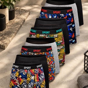 May include: A collection of nine boxer briefs arranged in a row, featuring various designs including solid black, solid gray, and multicolored patterns with graffiti-style graphics and the word SPORT printed on the elastic waistbands.
