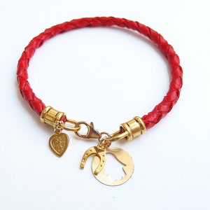 Puede incluir: Un brazalete de cuero trenzado rojo con abalorios dorados, incluyendo un corazón, una herradura y una campana.