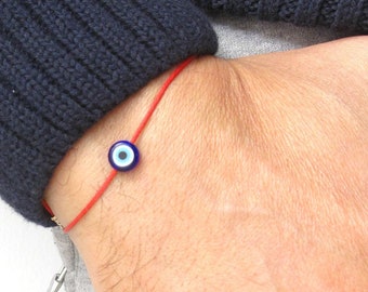 May include: A red string bracelet with a blue and white evil eye charm. The bracelet is worn on a person's wrist.
