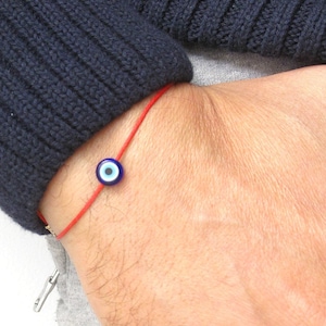 Può includere: Un braccialetto di corda rossa con un ciondolo di occhio turco blu e bianco. Il braccialetto è indossato al polso.