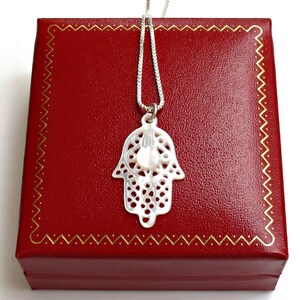 May include: A silver chain necklace with a white, openwork hamsa pendant. The pendant has a small, white pearl in the center. The necklace is displayed in a red velvet box.