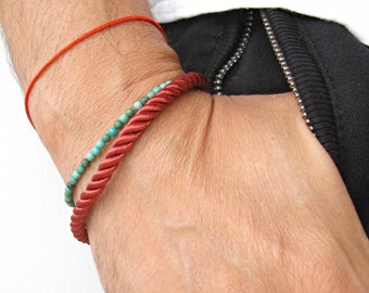 May include: Two bracelets on a wrist. One is a red rope bracelet, the other is a turquoise bead bracelet.