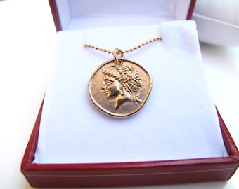 May include: A gold-colored pendant necklace featuring a detailed profile of a classical figure on a round coin-like charm. The necklace is presented in a red and white jewelry box, highlighting the pendant's intricate design and warm tones.