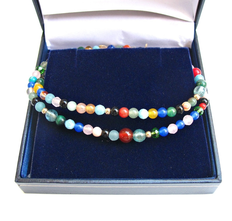 Pode incluir: Um colar de contas multicolorido com uma variedade de pedras, incluindo verde, azul, vermelho e amarelo, em uma caixa de joias de veludo azul.