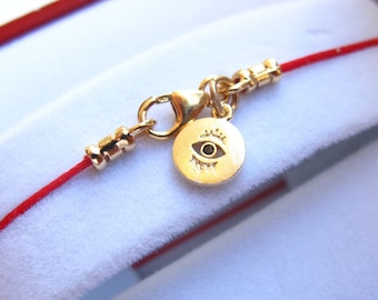 May include: A red string bracelet with a gold charm featuring an eye design. The charm has a small black stone in the center of the eye.