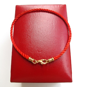 Puede incluir: Una pulsera de cordón rojo brillante con una textura de cuerda trenzada y un cierre de mosquetón de metal dorado. La pulsera tiene forma circular e incluye terminales de metal dorado que aseguran el cordón al mecanismo de cierre central.