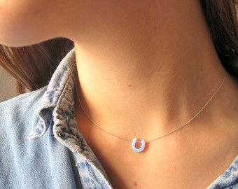 May include: A silver chain necklace with a light blue horseshoe pendant. The necklace is worn by a person wearing a denim shirt.