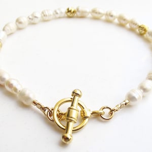 Puede incluir: Un brazalete de perlas blancas con un cierre de palanca dorado. El brazalete está hecho de pequeñas perlas redondas y tiene un acento dorado a cada lado del cierre.