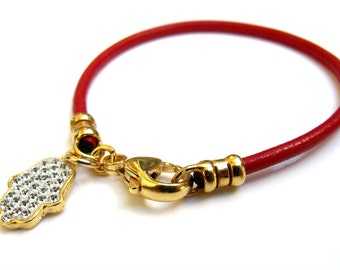 May include: A red leather bracelet with a gold clasp and a gold charm in the shape of a hamsa hand with white crystals.
