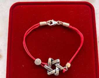 May include: A red string bracelet with a silver Star of David charm. The charm has Hebrew writing on it.
