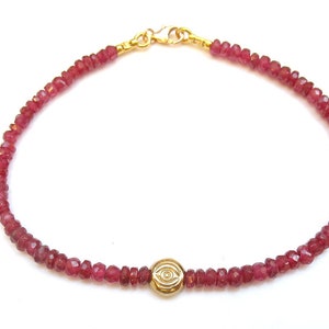 May include: A delicate bracelet featuring small, faceted, red beads strung together. A gold-colored charm with an eye design is centered on the bracelet. The clasp and connecting elements are also gold-colored.