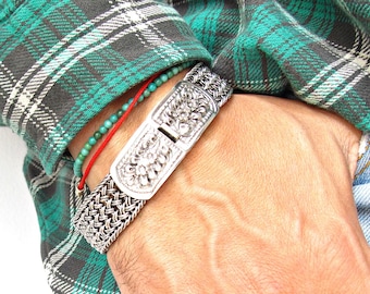 May include: A silver bracelet with a woven chain design and a decorative clasp. The bracelet is worn on a person's wrist with a turquoise and red beaded bracelet.