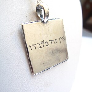 May include: A square silver-toned pendant hanging from a delicate silver chain. The pendant features a smooth, metallic surface with the Hebrew text "אין עוד מלבדו" engraved in black lettering across the center of the square face.