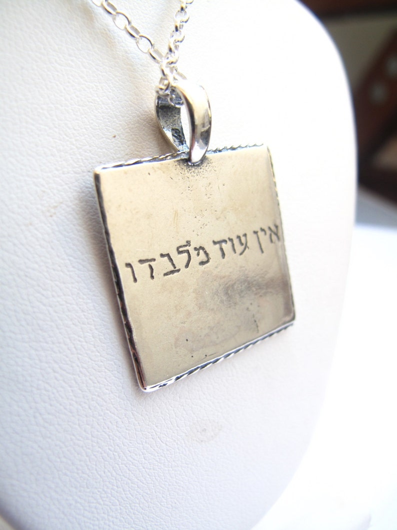 May include: A square silver-toned pendant hanging from a delicate silver chain. The pendant features a smooth, metallic surface with the Hebrew text "אין עוד מלבדו" engraved in black lettering across the center of the square face.