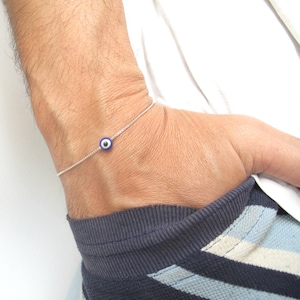 Puede incluir: Una delicada pulsera de cadena de plata con un pequeño dije redondo azul y blanco de ojo turco. La pulsera se lleva en la muñeca, mostrando el diseño minimalista y los colores vibrantes del dije. Es una pieza de joyería.