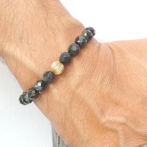 May include: A black beaded bracelet with a gold bead accent. The bracelet is made of faceted black beads and a single gold bead with a textured surface.