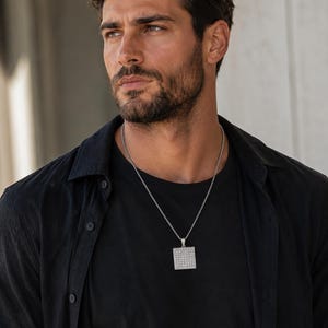 May include: A silver-toned square pendant necklace featuring a grid of small, reflective stones set into the metal surface. The pendant hangs from a thin, textured silver-toned chain worn over a plain black t-shirt and an unbuttoned black shirt.