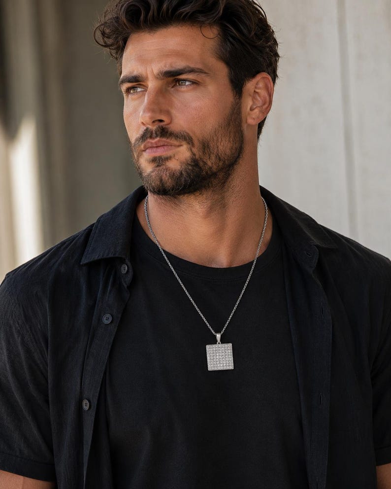 May include: A silver-toned square pendant necklace featuring a grid of small, reflective stones set into the metal surface. The pendant hangs from a thin, textured silver-toned chain worn over a plain black t-shirt and an unbuttoned black shirt.