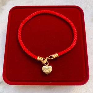 Pode incluir: Uma pulseira de cordão trançado vermelho com terminais de metal dourado e um pingente em forma de coração dourado. O pingente é gravado com um design de olho estilizado. A pulseira é fixada com um fecho lagosta de cor dourada.