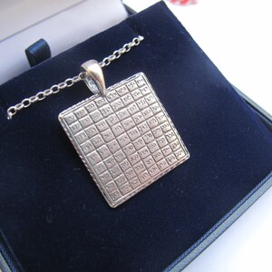 May include: A silver-toned square pendant featuring a grid of engraved Hebrew characters, suspended from a matching silver-toned chain. The pendant has a textured border and a polished bail, presenting a structured, geometric design.