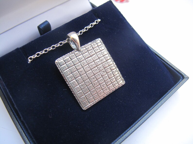 May include: A silver-toned square pendant featuring a grid of engraved Hebrew characters, suspended from a matching silver-toned chain. The pendant has a textured border and a polished bail, presenting a structured, geometric design.