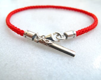 Pode incluir: Um bracelete de corda trançada vermelha com um fecho prateado e duas contas prateadas com o símbolo "++" gravado nelas.