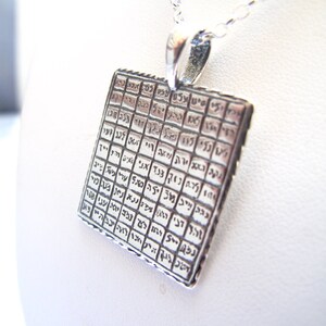 May include: A square silver pendant suspended from a matching silver chain. The pendant features a grid pattern containing engraved Hebrew characters within each individual square cell, creating a textured and detailed metallic surface design.