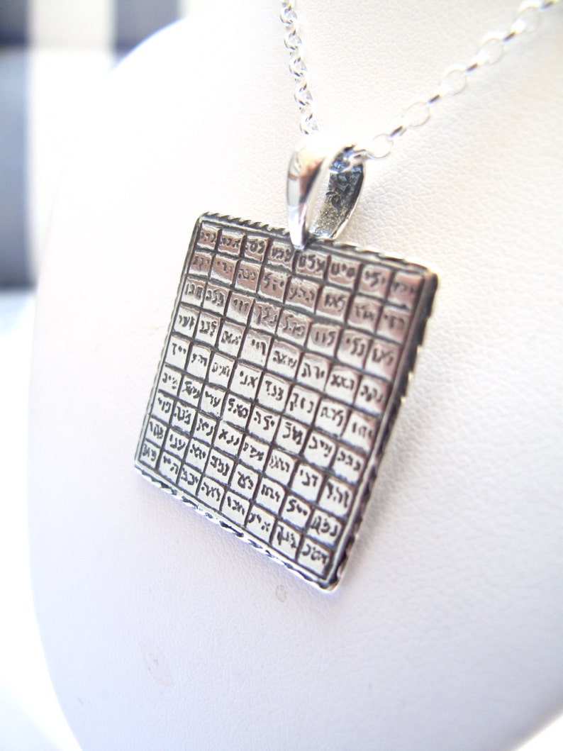May include: A square silver pendant suspended from a matching silver chain. The pendant features a grid pattern containing engraved Hebrew characters within each individual square cell, creating a textured and detailed metallic surface design.