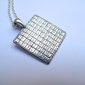 May include: A square silver pendant on a matching silver chain. The pendant features a grid pattern filled with engraved Hebrew characters in each small square section. The metal has a polished finish with darkened recessed areas to highlight the text.