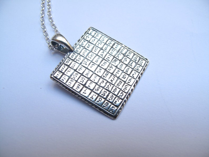 May include: A square silver pendant on a matching silver chain. The pendant features a grid pattern filled with engraved Hebrew characters in each small square section. The metal has a polished finish with darkened recessed areas to highlight the text.