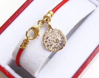 May include: A red leather bracelet with a gold-colored clasp and a decorative gold-colored pendant. The pendant has a circular shape with an openwork design. The bracelet is presented in a red and white jewelry box.