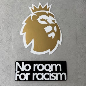 Puede incluir: Una calcomanía que presenta un icono de cabeza de león dorado con un contorno blanco y una corona en la parte superior. Debajo del león hay una pegatina rectangular negra con el texto blanco "No room for racism" en fuente sans-serif.