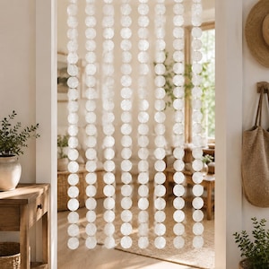 May include: A hanging room divider curtain composed of multiple vertical strands of translucent, circular white shell discs. The discs are linked together in a repeating pattern, creating a semi-sheer, shimmering partition that hangs from a top rod.