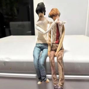 Könnte beinhalten: Zwei Stoffpuppen sitzen nebeneinander. Die linke Puppe hat schwarze Haare, ein weißes Langarmshirt und eine blaue Jeans. Die rechte Puppe hat braune Haare, ein weißes Hemd mit roter Krawatte und eine braune Hose. Ein Arm liegt um die rechte Puppe.