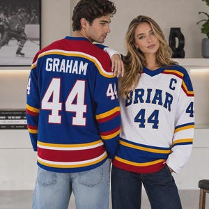 Peut inclure: Un maillot de hockey bleu avec le nom GRAHAM et le numéro 44 en blanc, et un maillot de hockey blanc avec le nom BRIAR, le numéro 44 et un emblème C en bleu. Les deux maillots présentent des rayures horizontales rouges et jaunes.