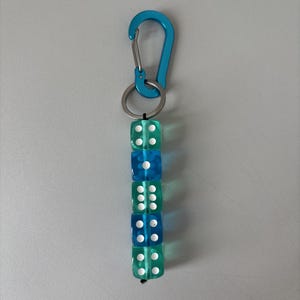 Könnte beinhalten: Ein Schlüsselanhänger mit einem leuchtend blauen Metallkarabiner an einem silbernen Schlüsselring. Daran hängt eine vertikale Reihe aus fünf durchsichtigen, quadratischen Würfeln in abwechselndem Türkis und Blau, die mit weißen Punkten versehen sind.