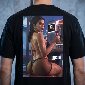 May include: A black t-shirt featuring a rectangular graphic print on the back. The print depicts a person with dark hair wearing a black bikini and fishnet bottoms, holding a glass. The Rockstar Games logo and the text LUCIA are visible.