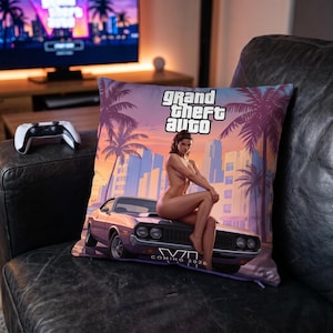 Pode incluir: Uma almofada quadrada com uma estampa gráfica de Grand Theft Auto. O design mostra uma pessoa sentada no capô de um carro esportivo roxo contra um cenário urbano ao pôr do sol com palmeiras. O texto diz Grand Theft Auto e Coming 2026.